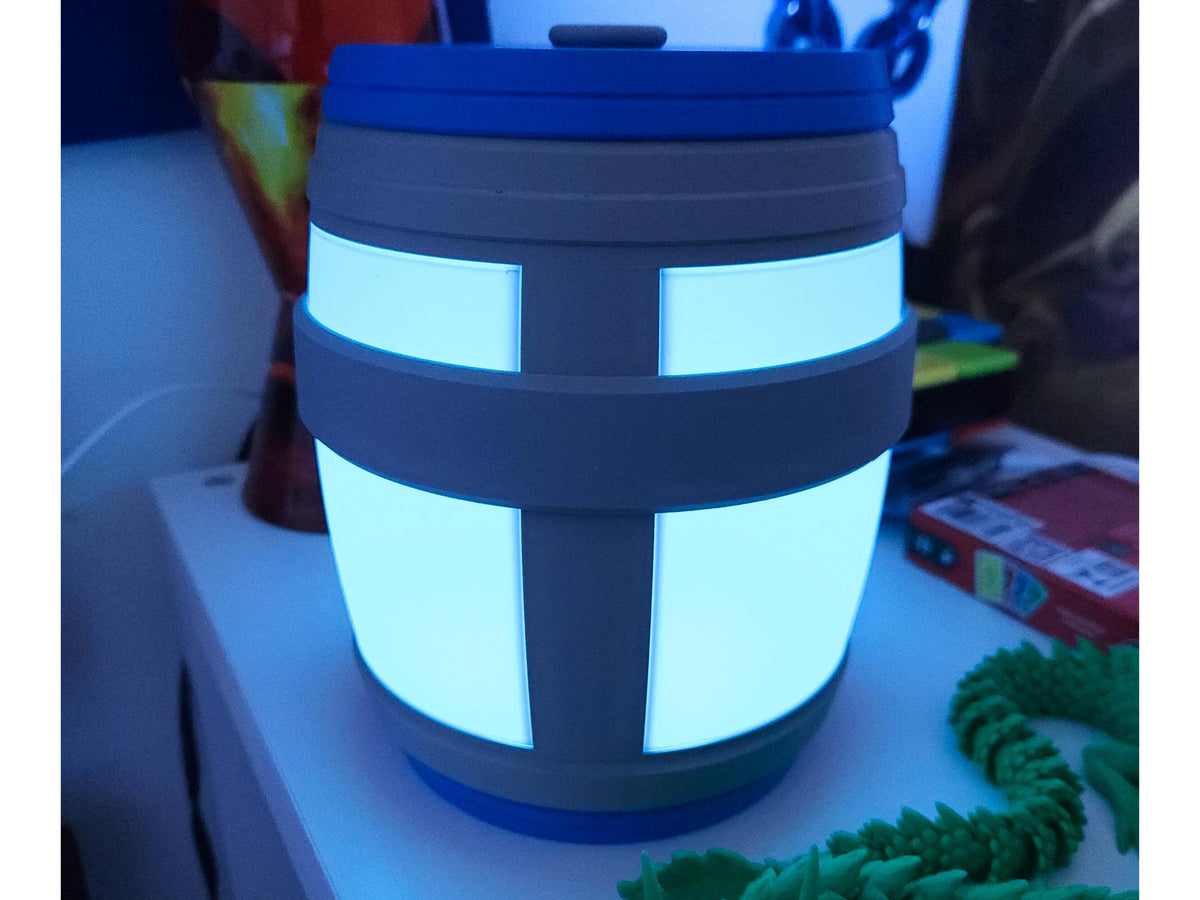 A glowing blue barrel-shaped LED lamp with a gray band design, inspired by gaming aesthetics. The lamp emits a soft ambient light, making it perfect for nightstands or gaming setups.