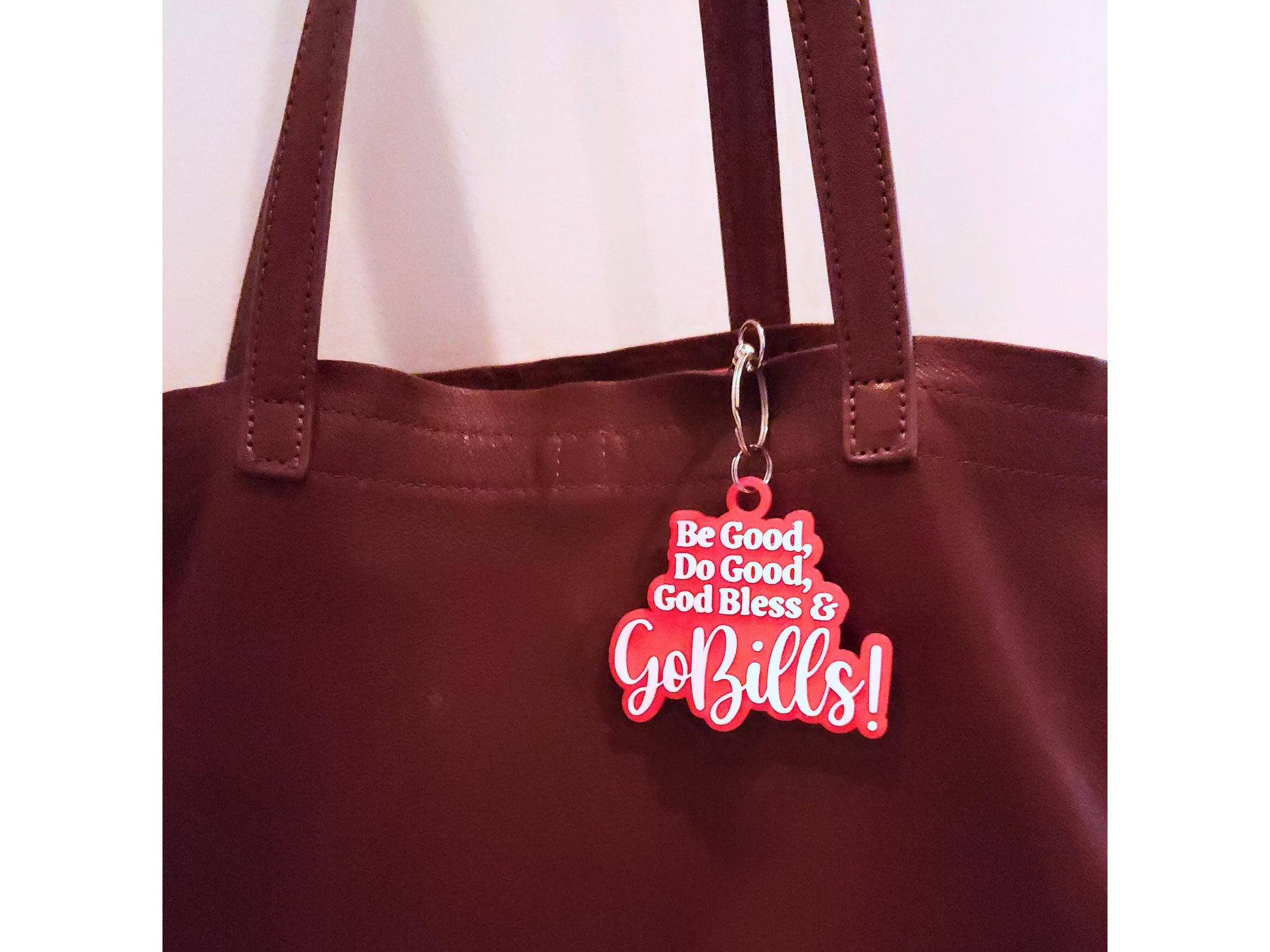 A red keychain displayed on a handbag. The keychain says Be Good, Do Good, God Bless, Go bills.