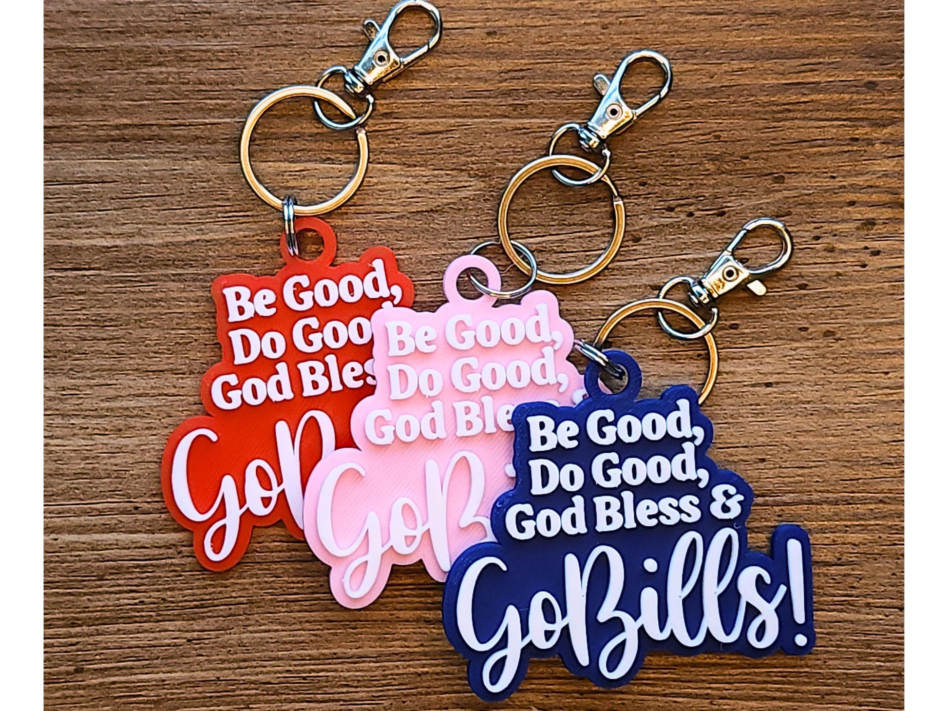 A set of 3 keychains that say Be Good, Do Good, God Bless, Go Bills in white letters on a background that is pink, blue, or red.