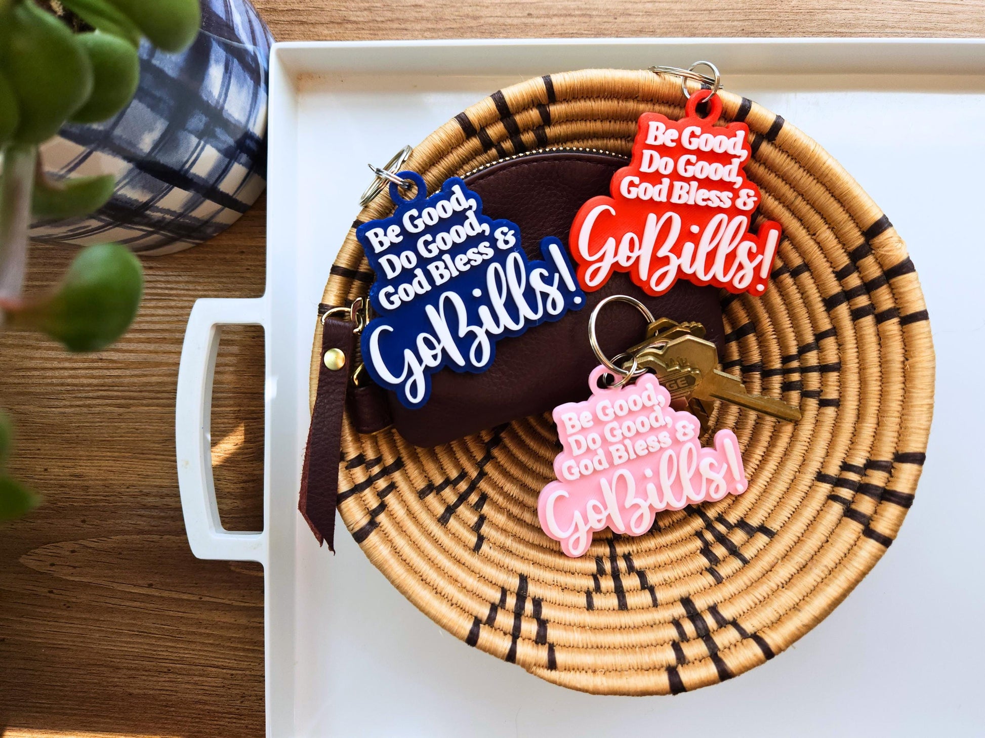 3 buffalo bills keychains in a basket, each says Be Good Do good God Bless Go Bills in honor of Josh Allen&#39;s MVP speech. One is red, one is blue, one is pink and each has white letters.