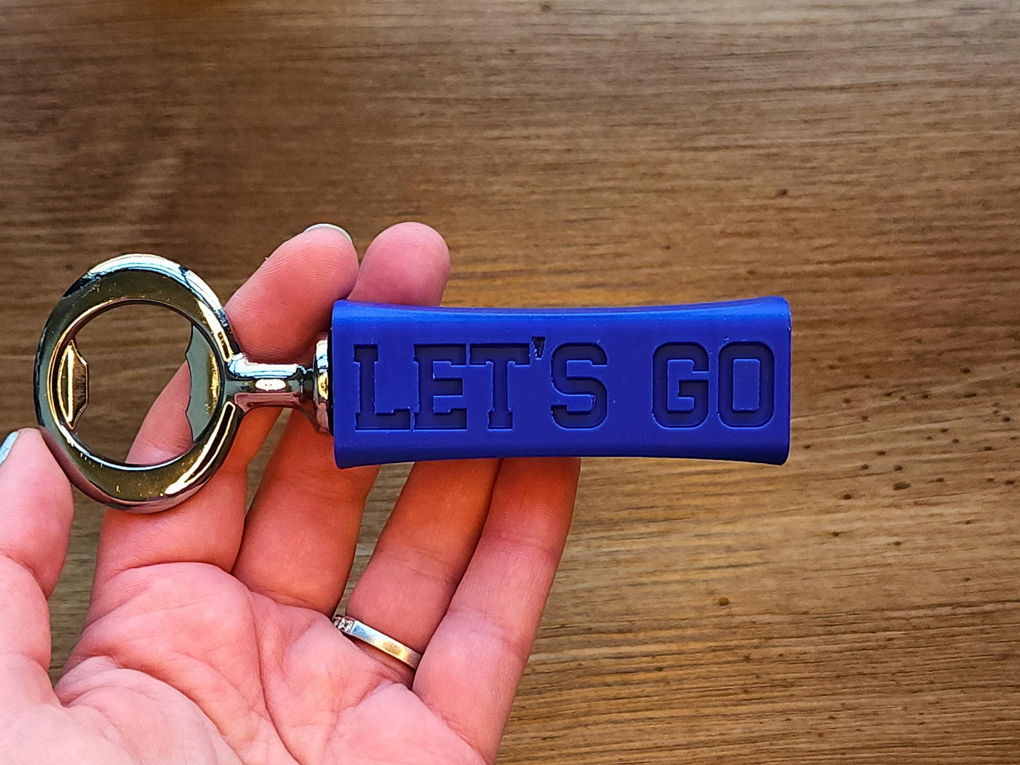 Close-up of a hand holding the bottle opener, showing the text Let’s Go clearly embossed on the grip.