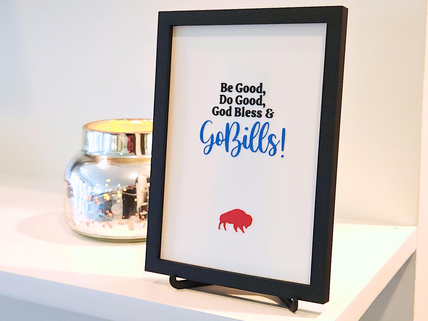A close-up side perspective of the same 3D printed sign, showing the layered texture of the text and buffalo icon. The frame and attached stand give it a clean, sturdy look. Light from the left creates soft shadows that highlight the 3D print&#39;s depth and finish. Decorative candle holder adds a reflective accent beside it.