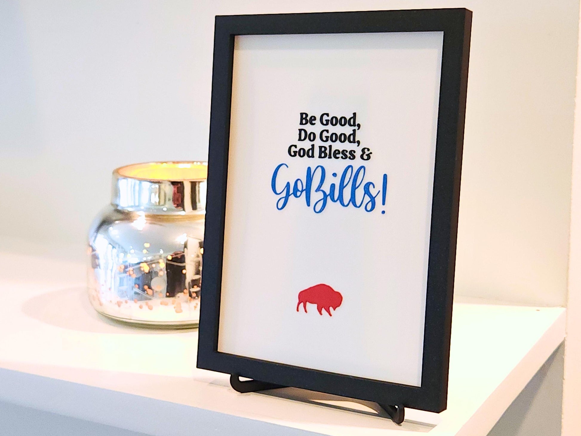 A close-up side perspective of the same 3D printed sign, showing the layered texture of the text and buffalo icon. The frame and attached stand give it a clean, sturdy look. Light from the left creates soft shadows that highlight the 3D print&#39;s depth and finish. Decorative candle holder adds a reflective accent beside it.