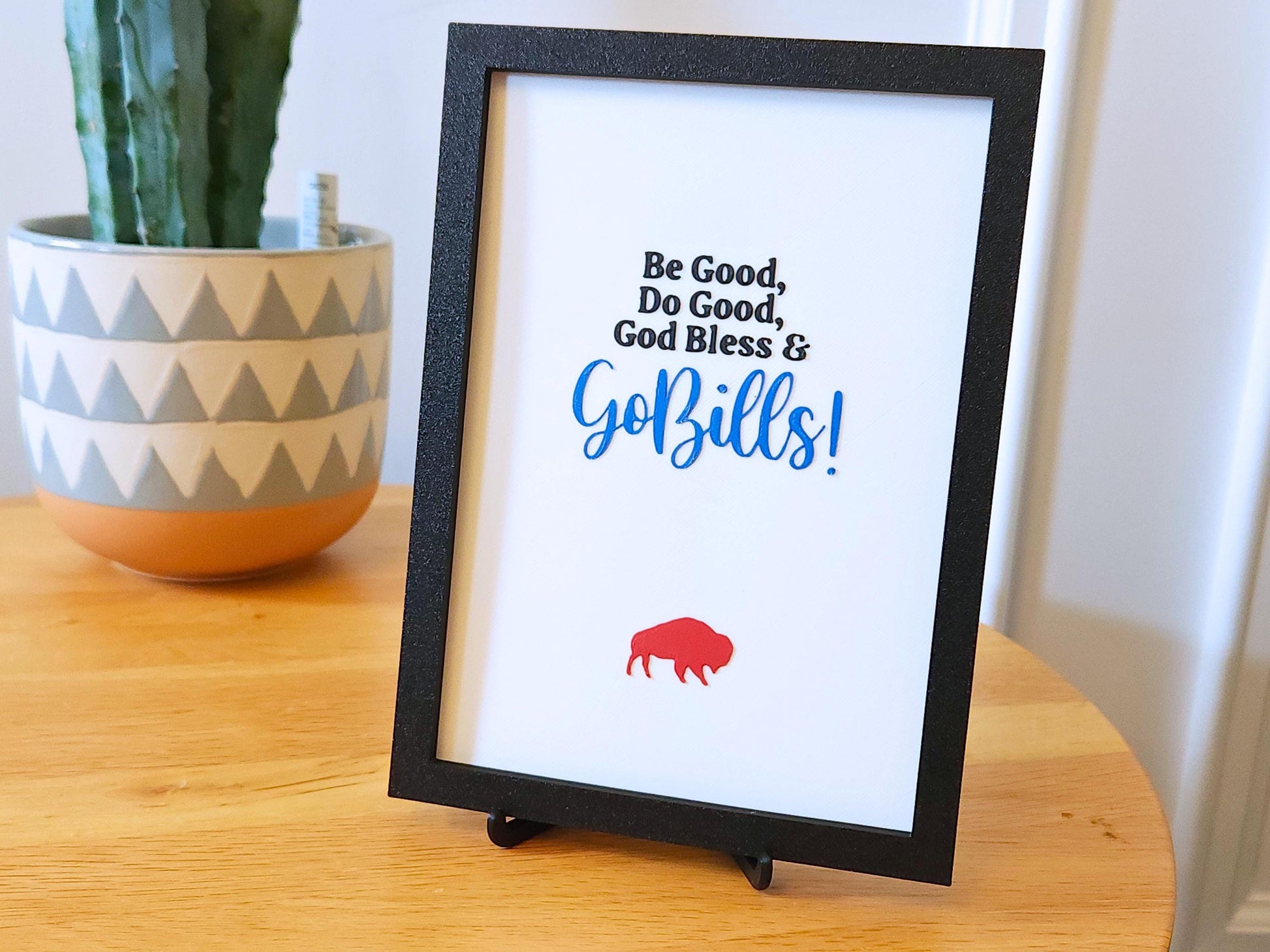 A bright, daylight photo of the 3D printed Buffalo-themed sign from a slightly lower angle. The lighting highlights the smooth surface of the print and gives depth to the framed design. The black easel stand supports the sign, showing it&#39;s freestanding. The red buffalo icon at the bottom balances the bold script above it, making the overall composition eye-catching and full of team spirit.