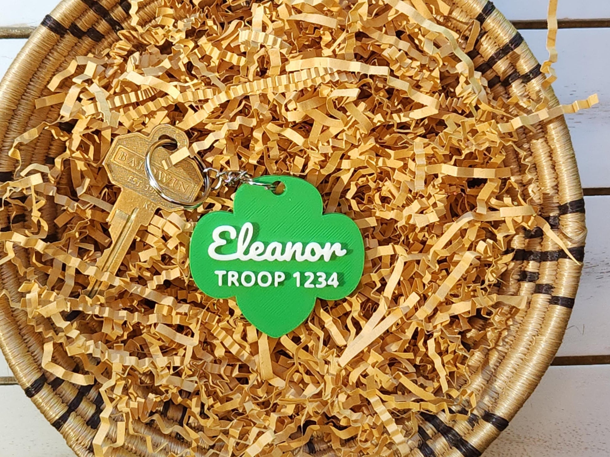 Girl Scout gift idea: green trefoil keychain with name and troop number nestled in shredded paper for packaging, perfect for end-of-year troop gifts or bridging ceremonies.