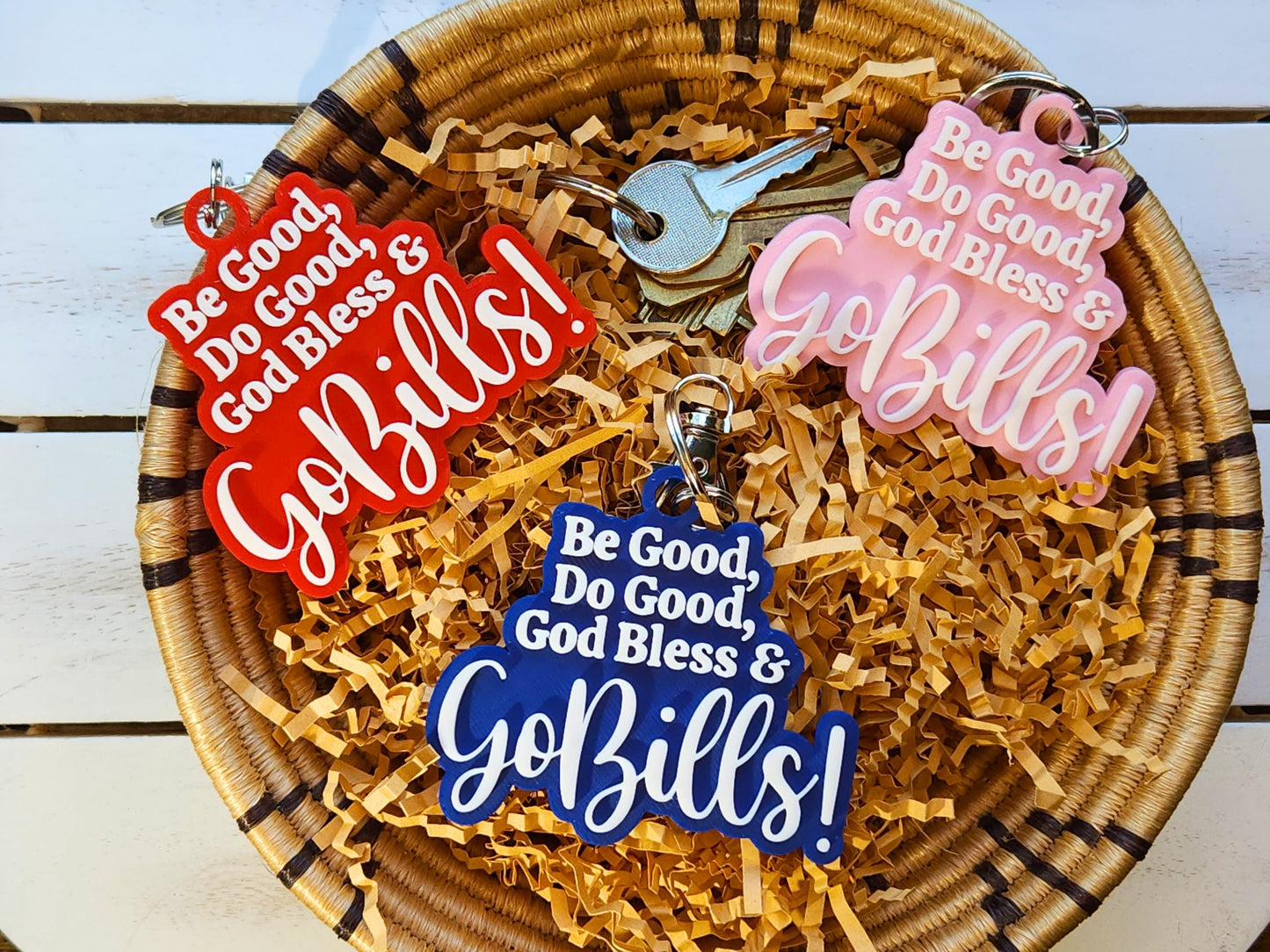 3 buffalo bills keychains in a basket, each says Be Good Do good God Bless Go Bills in honor of Josh Allen&#39;s MVP speech. One is red, one is blue, one is pink and each has white letters.