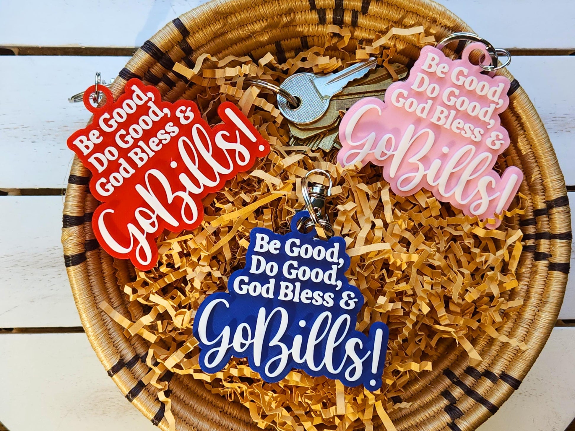3 buffalo bills keychains in a basket, each says Be Good Do good God Bless Go Bills in honor of Josh Allen&#39;s MVP speech. One is red, one is blue, one is pink and each has white letters.