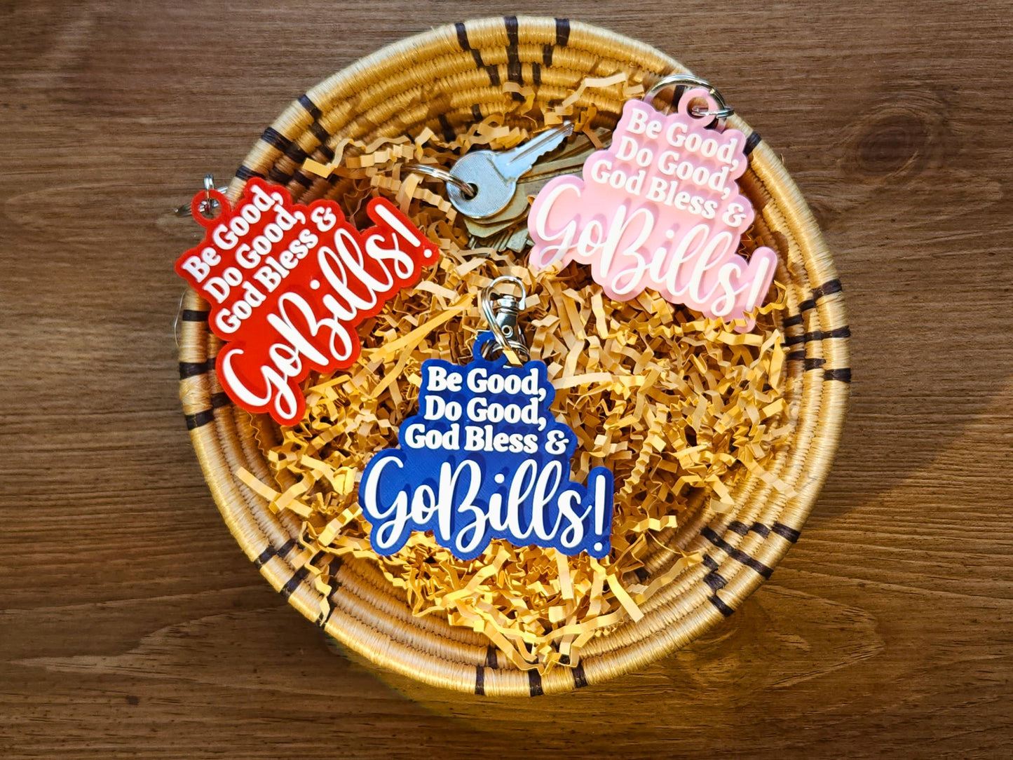 3 buffalo bills keychains in a basket, each says Be Good Do good God Bless Go Bills in honor of Josh Allen&#39;s MVP speech. One is red, one is blue, one is pink and each has white letters.