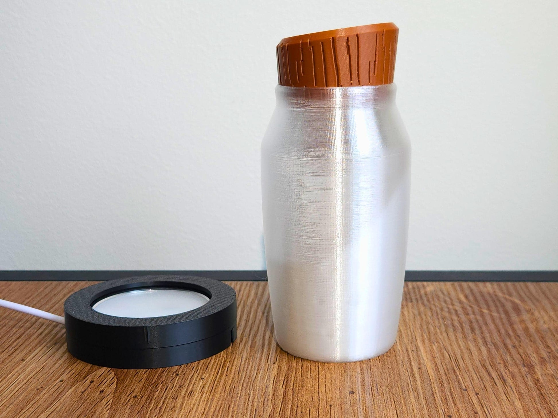 A potion bottle with a brown wood-textured cap standing next to a black LED light base, photographed on a wooden tabletop with a white wall background.