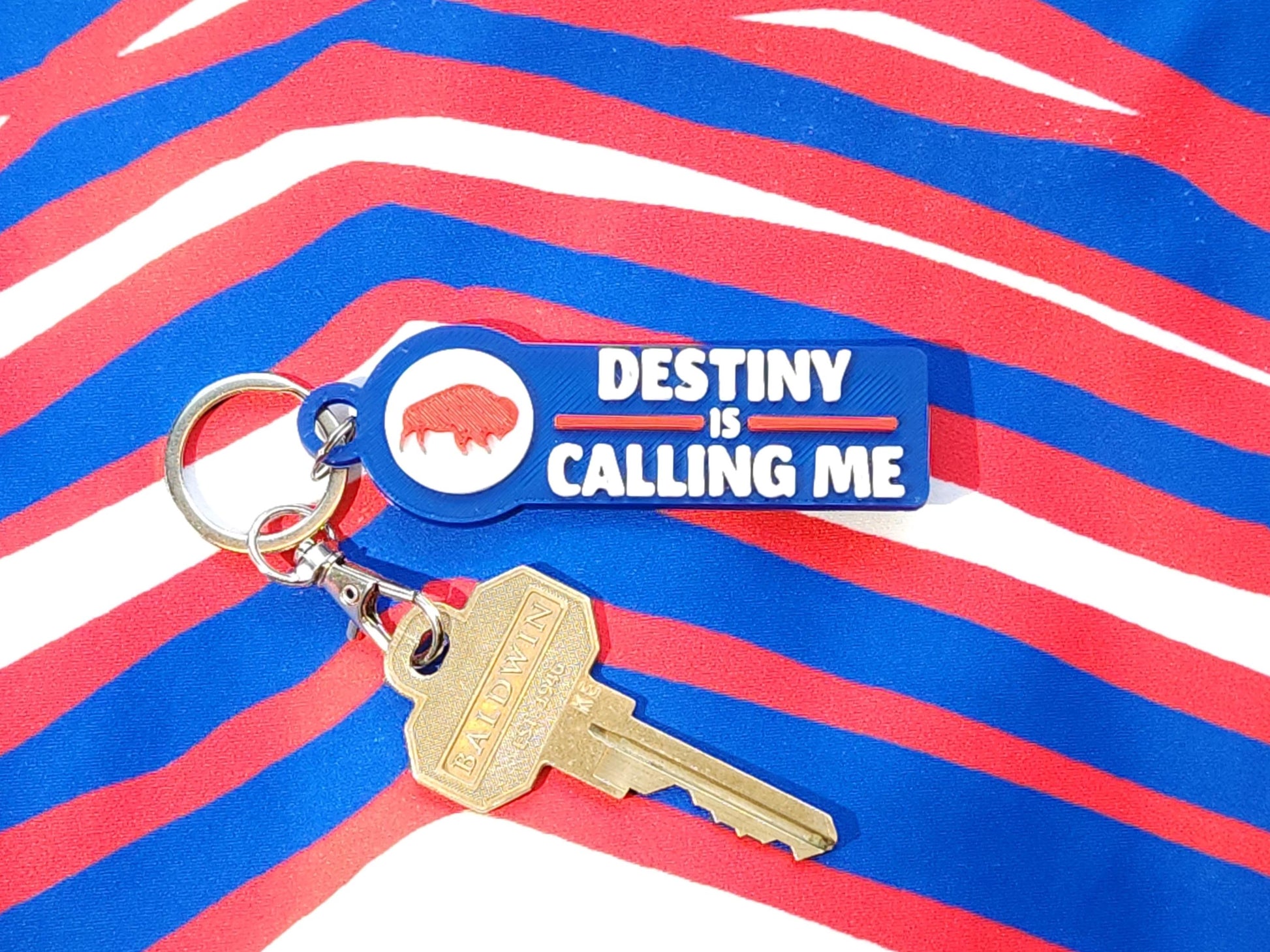 A blue keychain that says Destiny is Calling Me Go Bills displayed on red white and blue zubaz background.