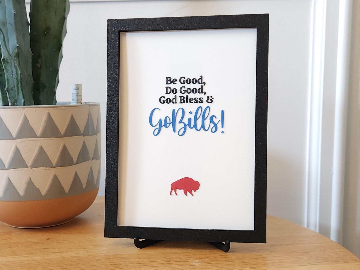 The sign is displayed in a casual setting on a light wood surface with a tall green cactus in a geometric-patterned terracotta and cream pot to the left. The vibrant red buffalo and Bills blue script stand out against the crisp white background, and the frame’s black texture is clearly visible. The photo emphasizes the sign’s compact size and how well it fits into a modern, minimalist space.