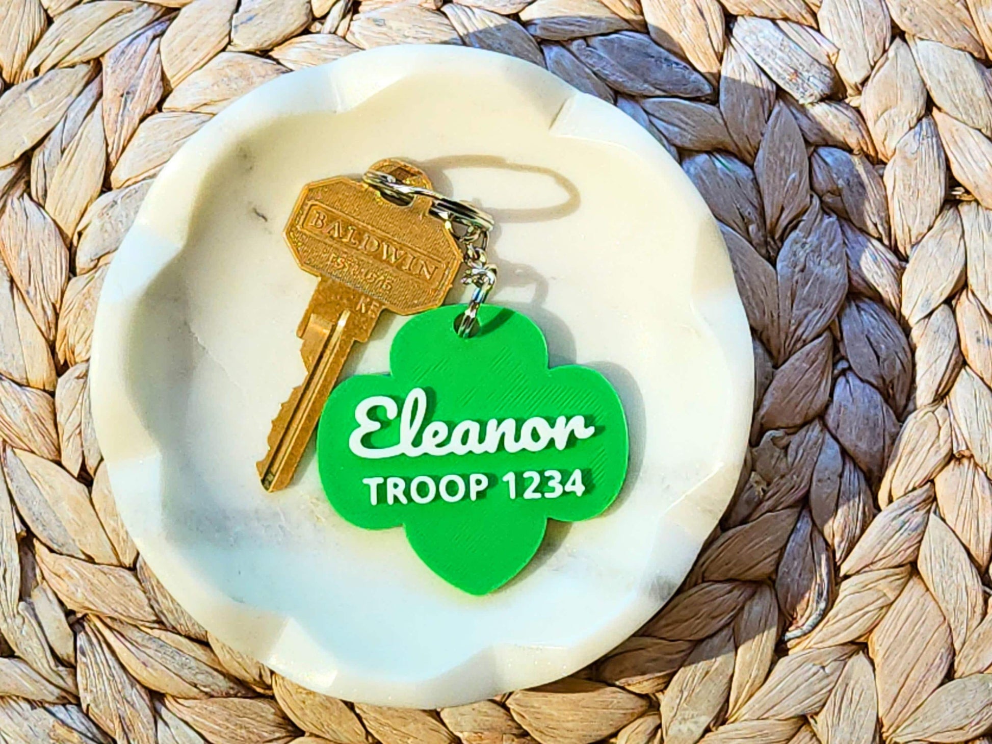 Personalized Girl Scout keychain with green trefoil shape, featuring name &quot;Eleanor&quot; and troop number 1234, attached to a gold key and displayed in a white dish on a woven mat.