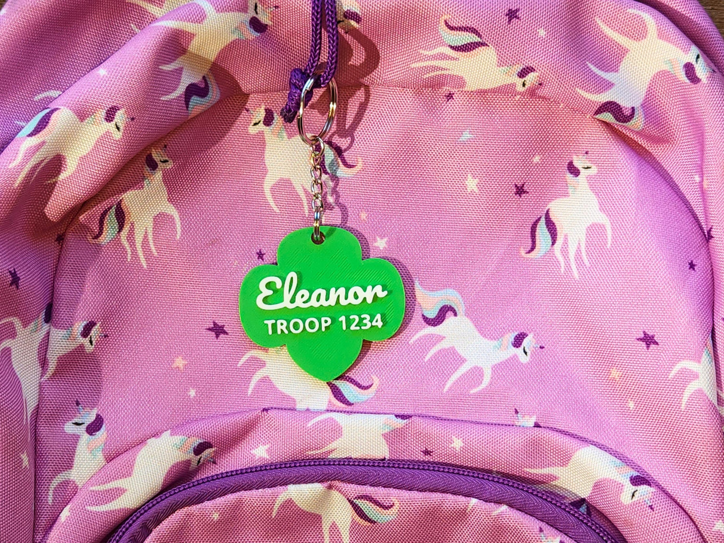 Cute personalized Girl Scout backpack keychain for kids, shown on a pink unicorn-themed backpack with green trefoil tag reading &quot;Eleanor - Troop 1234.&quot;