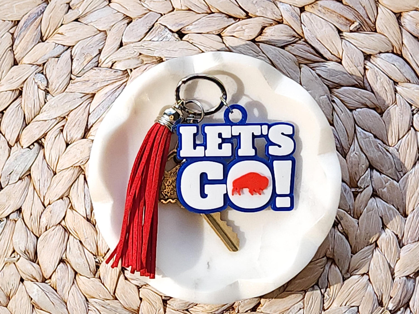 View of Let&#39;s Go Buffalo football keychain with optional red tassel in a white dish on a woven background.