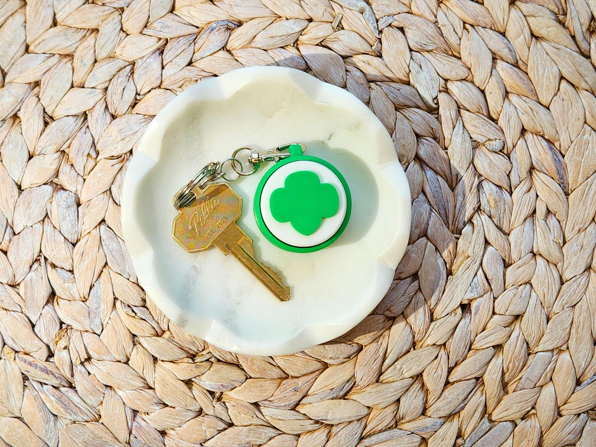 A green and white Girl Scout-themed fidget keychain with a trefoil design, resting in a marble dish beside a key.