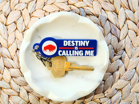 View of a blue keychain that says Destiny is Calling me Go Bills in a small white bowl with a key attached.