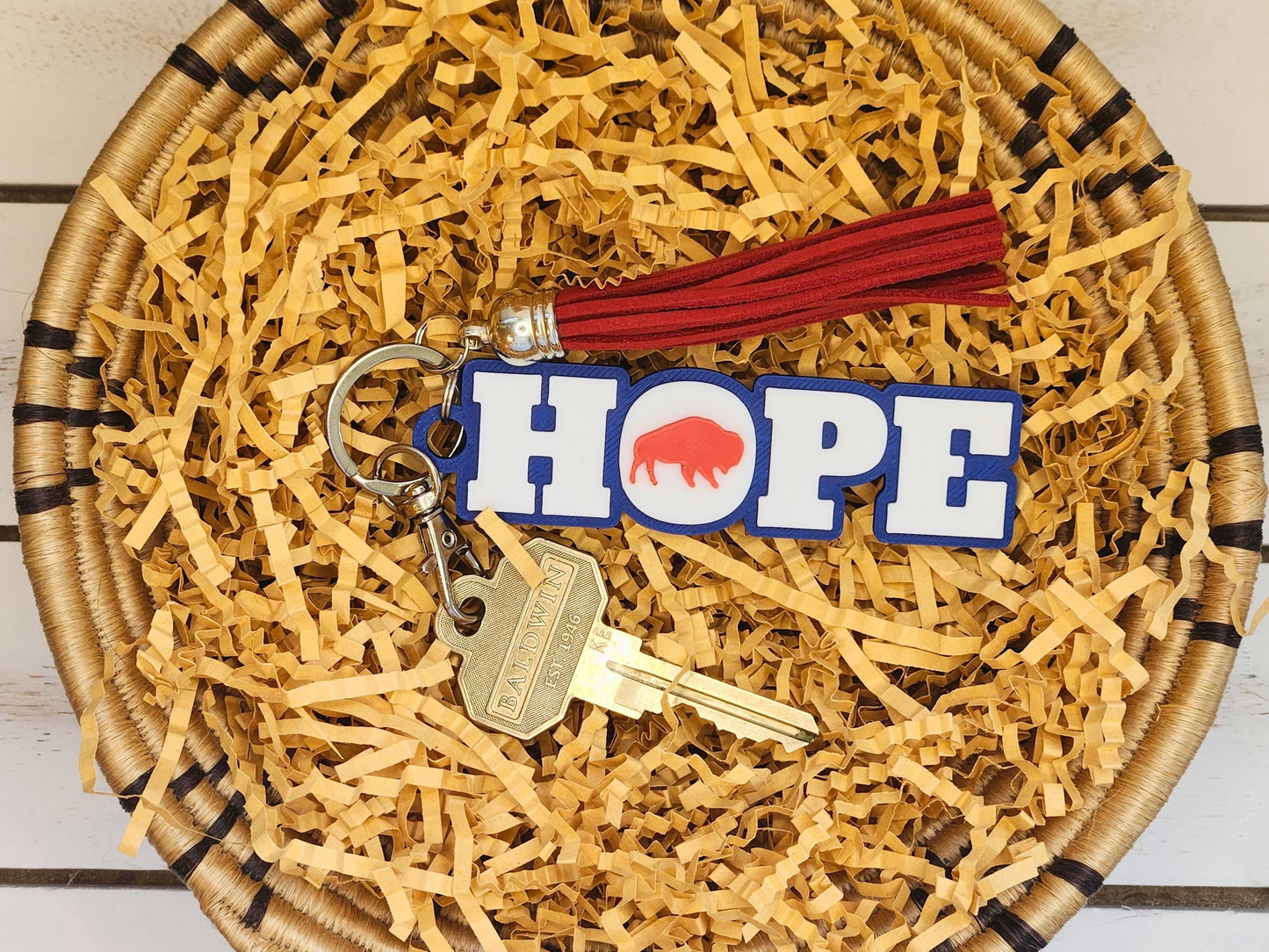 The “HOPE” keychain with a red buffalo icon in place of the “O,” shown in a woven basket with crinkle paper, attached to a brass key and a red tassel.