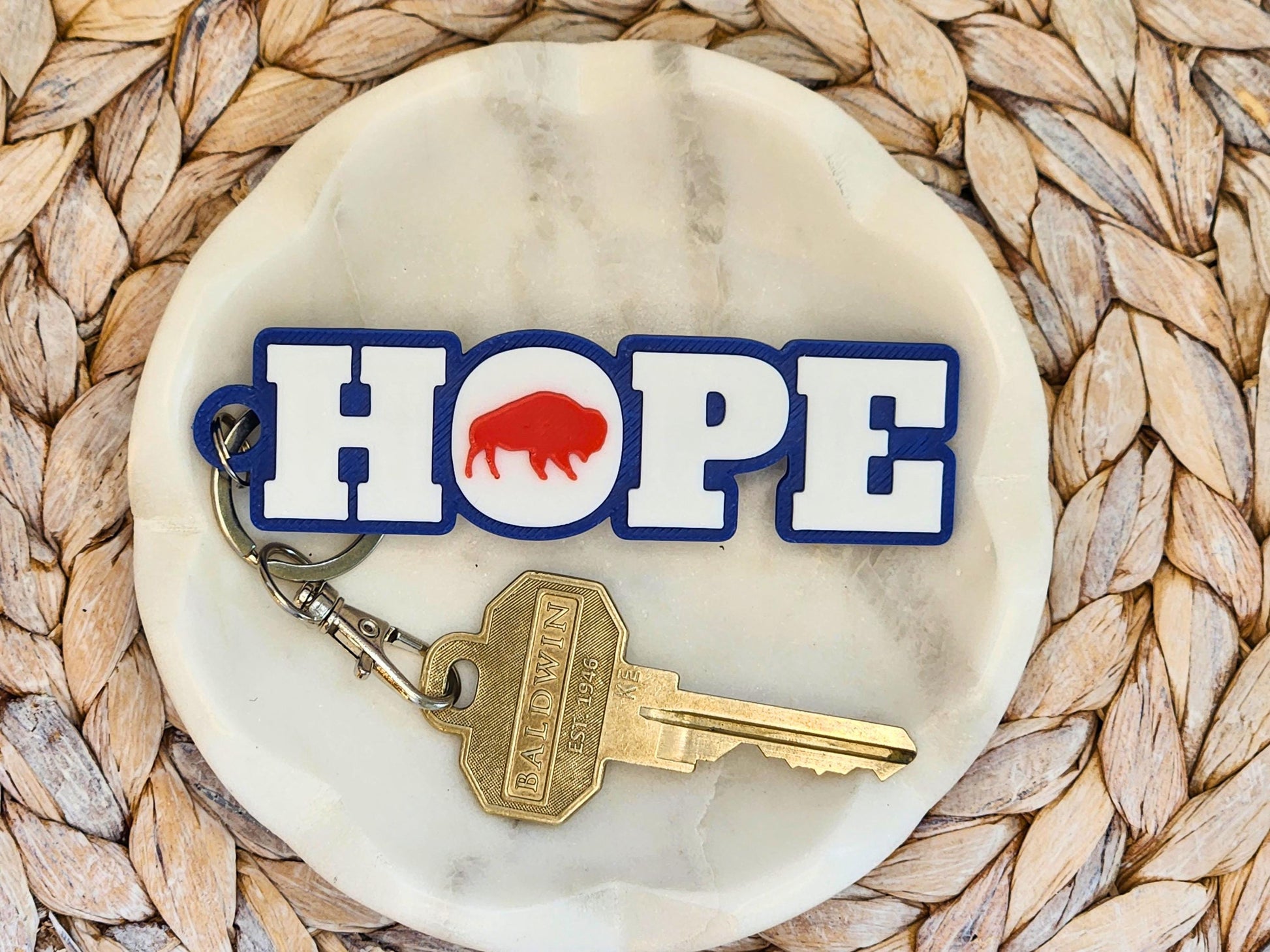 Buffalo football keychain with HOPE design, featuring red buffalo silhouette on white and blue lettering, displayed on a marble dish with a brass key.