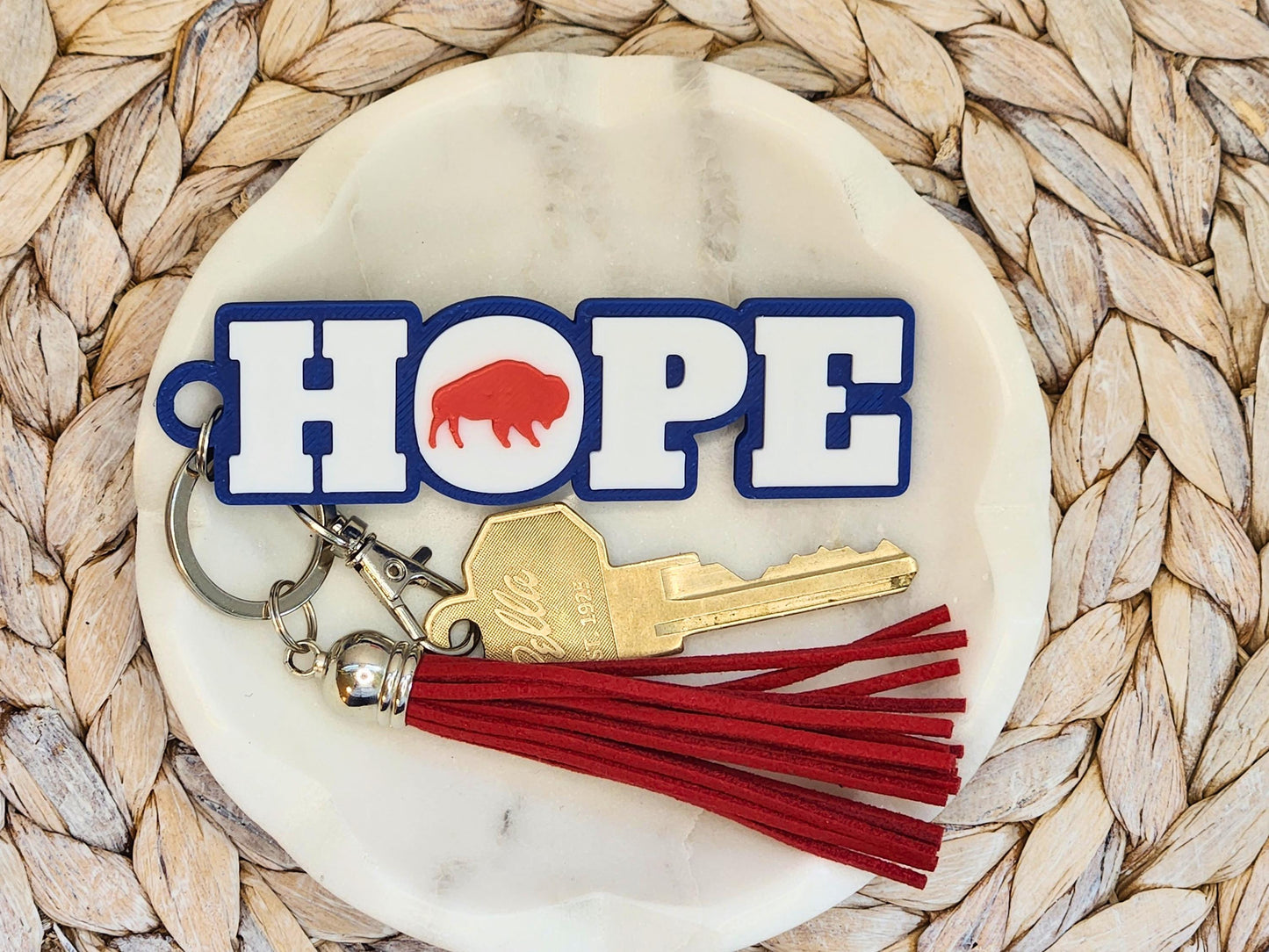 The Buffalo football keychain shown again on a marble dish with woven background, featuring the red tassel and the word &quot;Hope&quot; and brass key clearly displayed.