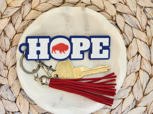 The Buffalo football keychain shown again on a marble dish with woven background, featuring the red tassel and the word &quot;Hope&quot; and brass key clearly displayed.