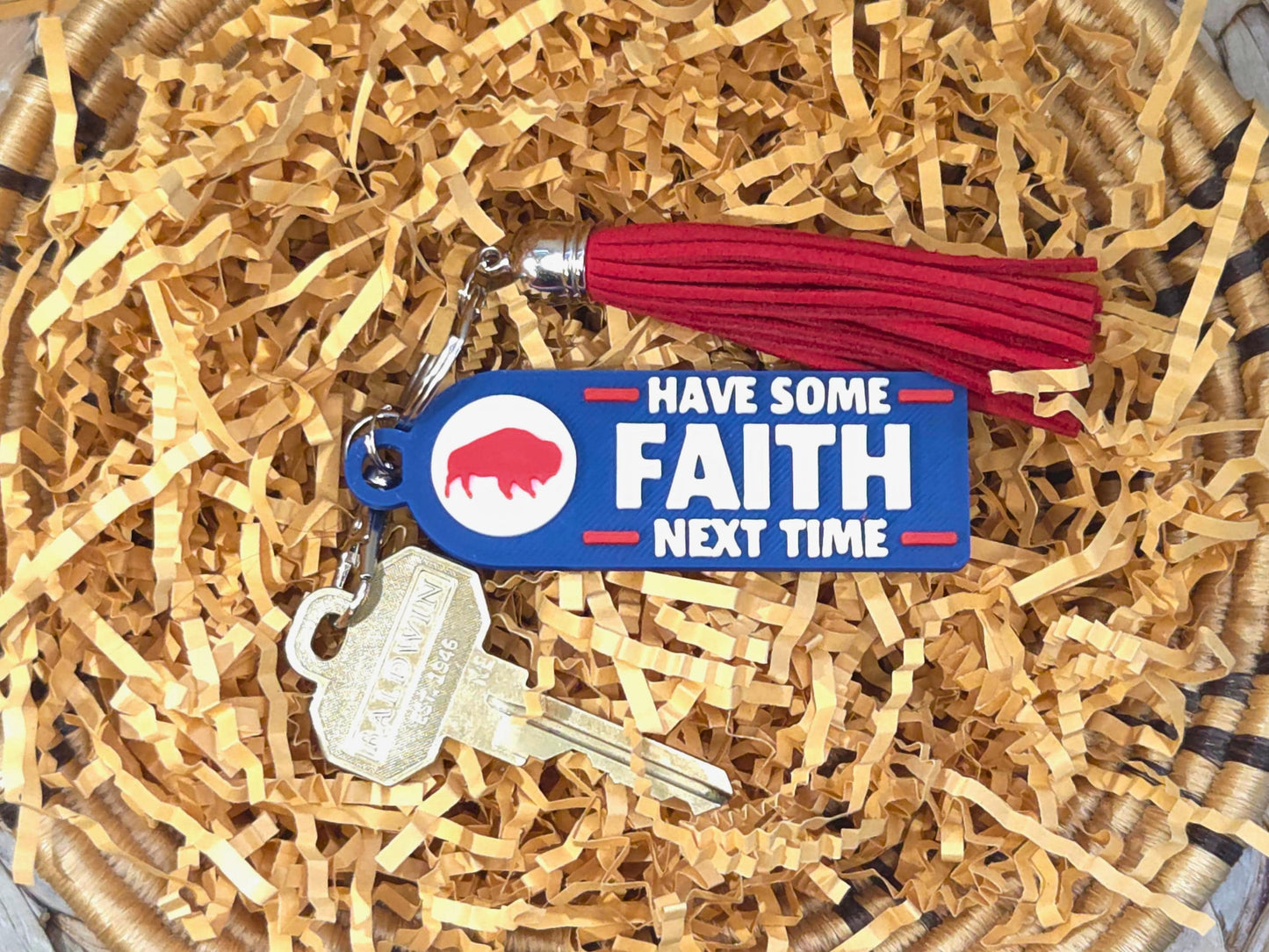 Blue and red 3D printed keychain with the phrase &#39;Have Some Faith Next Time&#39; attached to gold keys and a red tassel, displayed in a basket with crinkle paper.