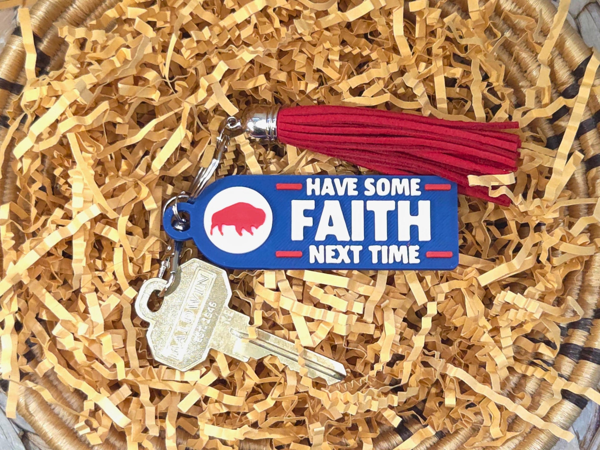 Blue and red 3D printed keychain with the phrase &#39;Have Some Faith Next Time&#39; attached to gold keys and a red tassel, displayed in a basket with crinkle paper.