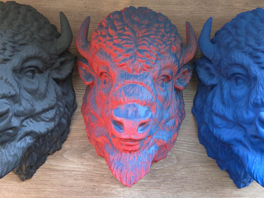 View of 3 3D printed bison heads in black, red/blue, and blue pictured side by side on a wooden background.