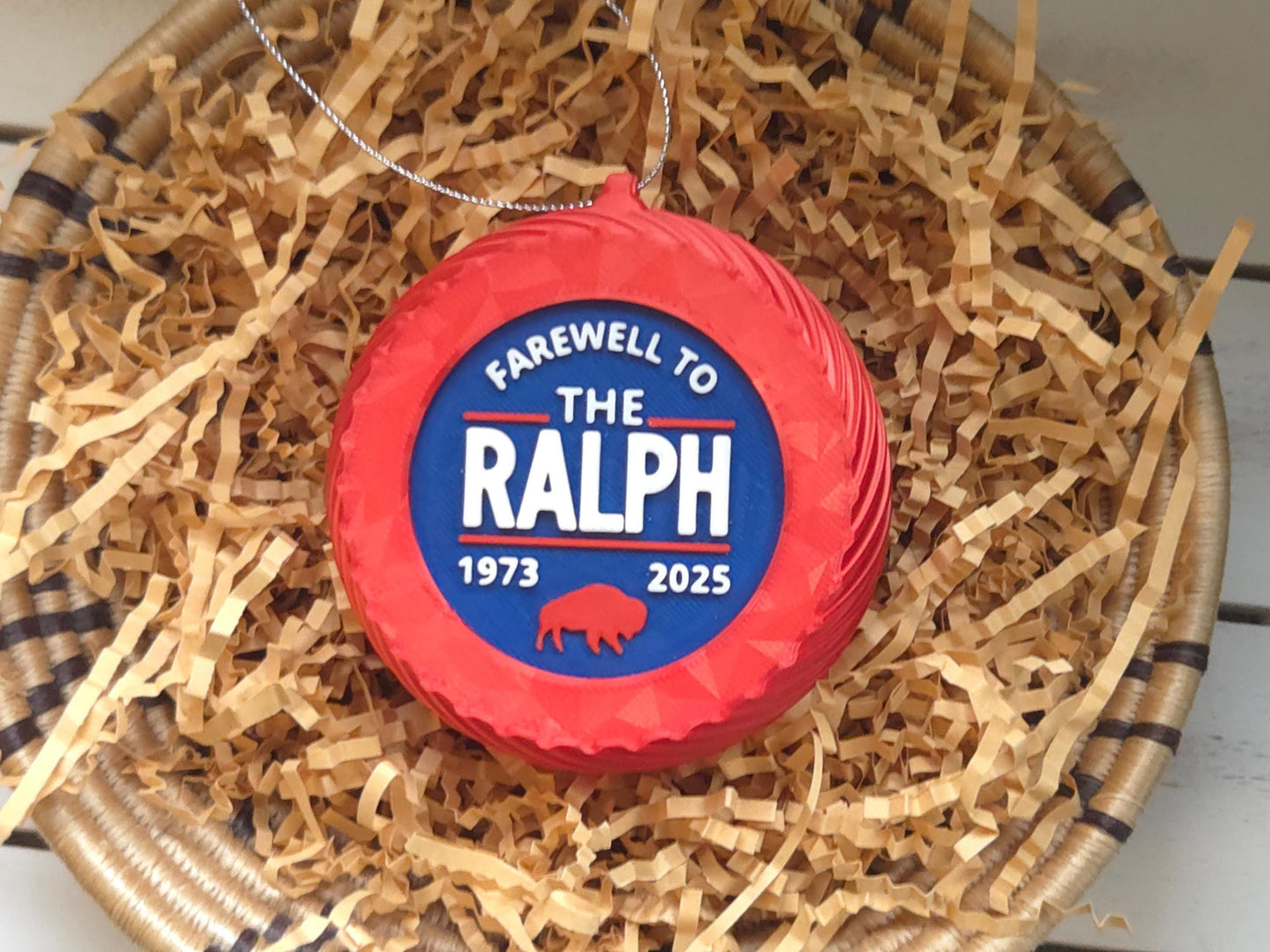 A 4" tall blue Christmas ornament with the words “Farewell to The Ralph 1973–2025” in bold white letters, a red buffalo graphic, and a silver hanging loop. Displayed on a bed of crinkled kraft paper