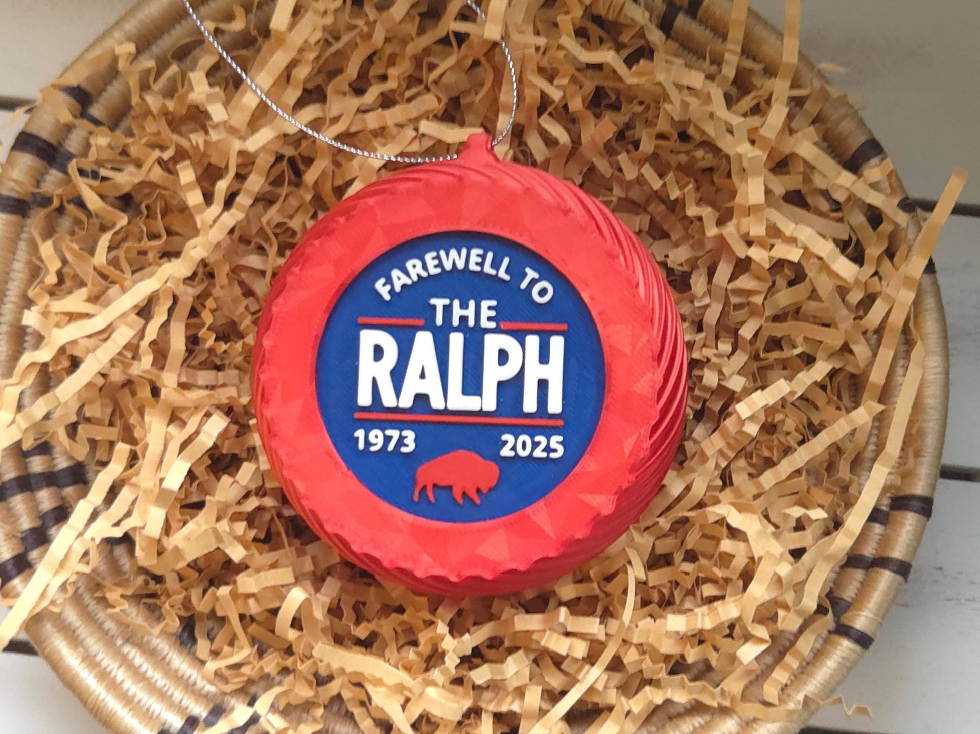 A 4" tall blue Christmas ornament with the words “Farewell to The Ralph 1973–2025” in bold white letters, a red buffalo graphic, and a silver hanging loop. Displayed on a bed of crinkled kraft paper