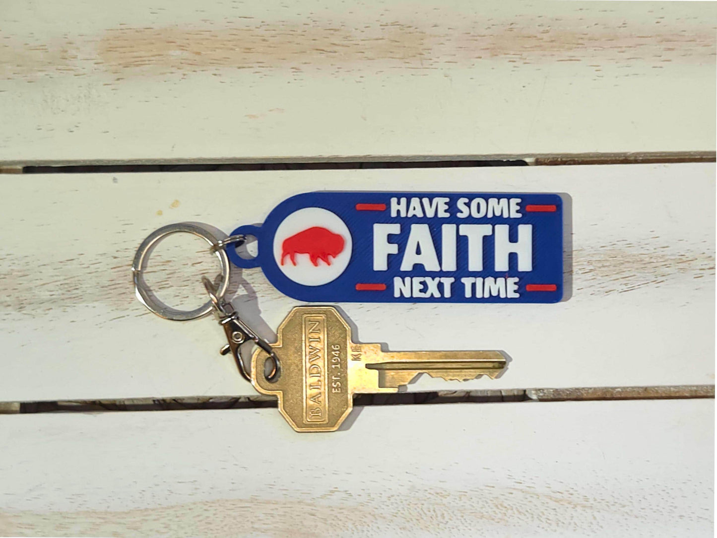 Buffalo-themed blue keychain with white and red text reading &#39;Have Some Faith Next Time,&#39; attached to gold keys, shown on a white wooden surface.