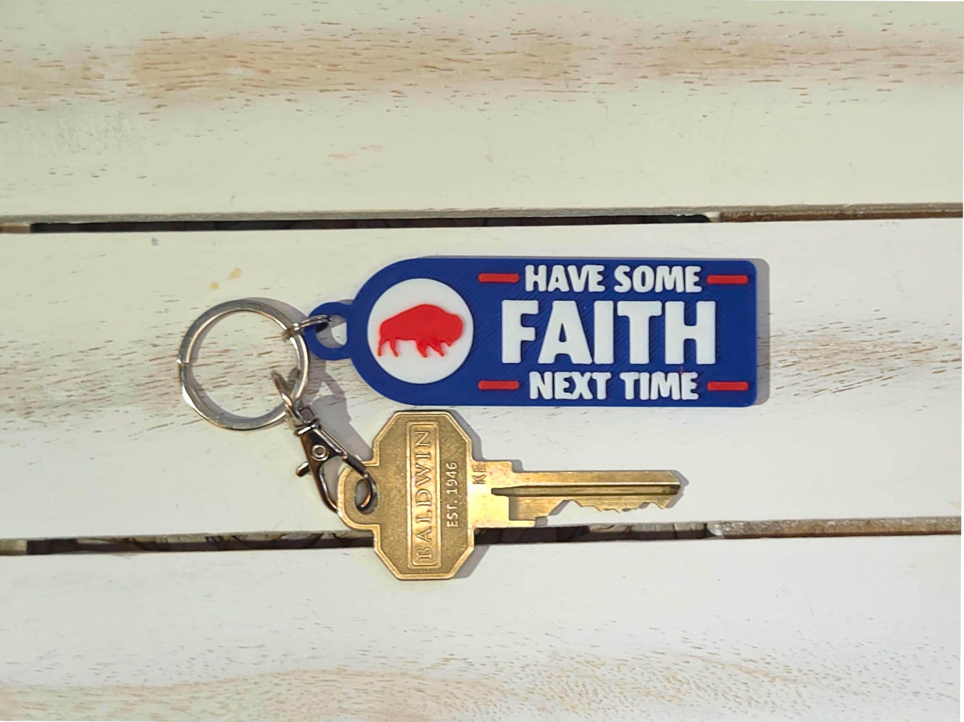 Buffalo-themed blue keychain with white and red text reading &#39;Have Some Faith Next Time,&#39; attached to gold keys, shown on a white wooden surface.