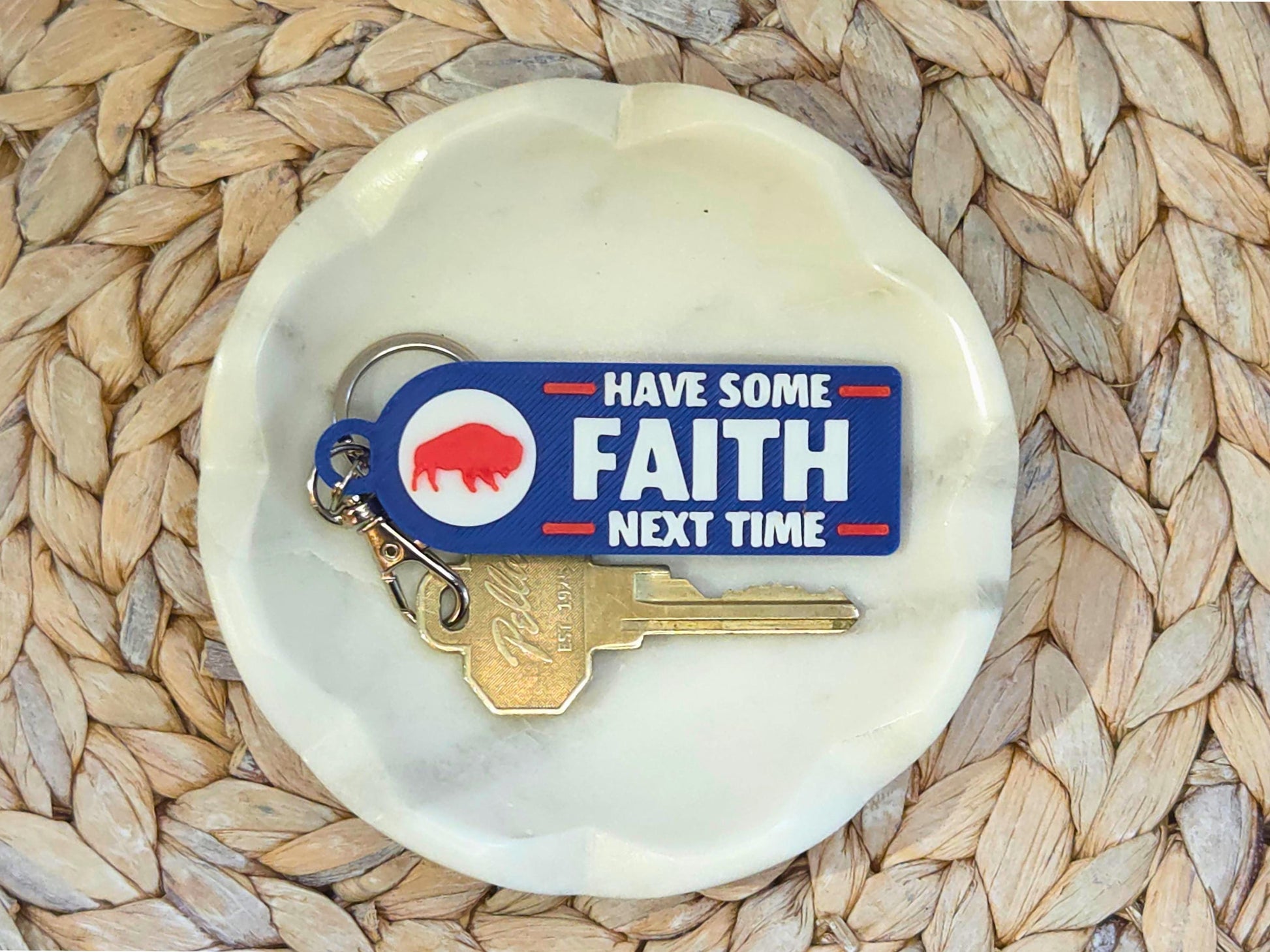 Keychain with bold white text &#39;Have Some Faith Next Time&#39; on blue background, with red buffalo silhouette, placed on a small white dish over woven mat