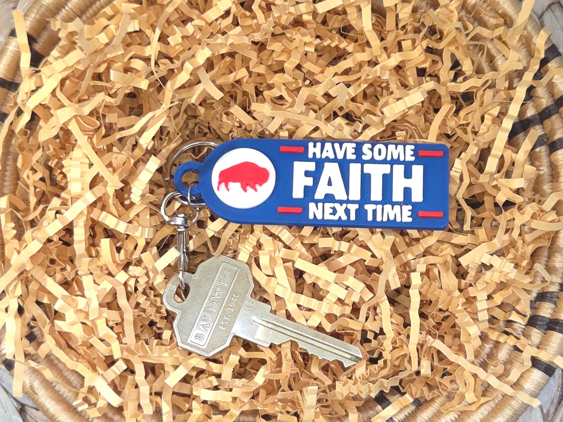 Buffalo-inspired keychain on shredded paper filler, showing the words &#39;Have Some Faith Next Time&#39; in white and red on blue plastic