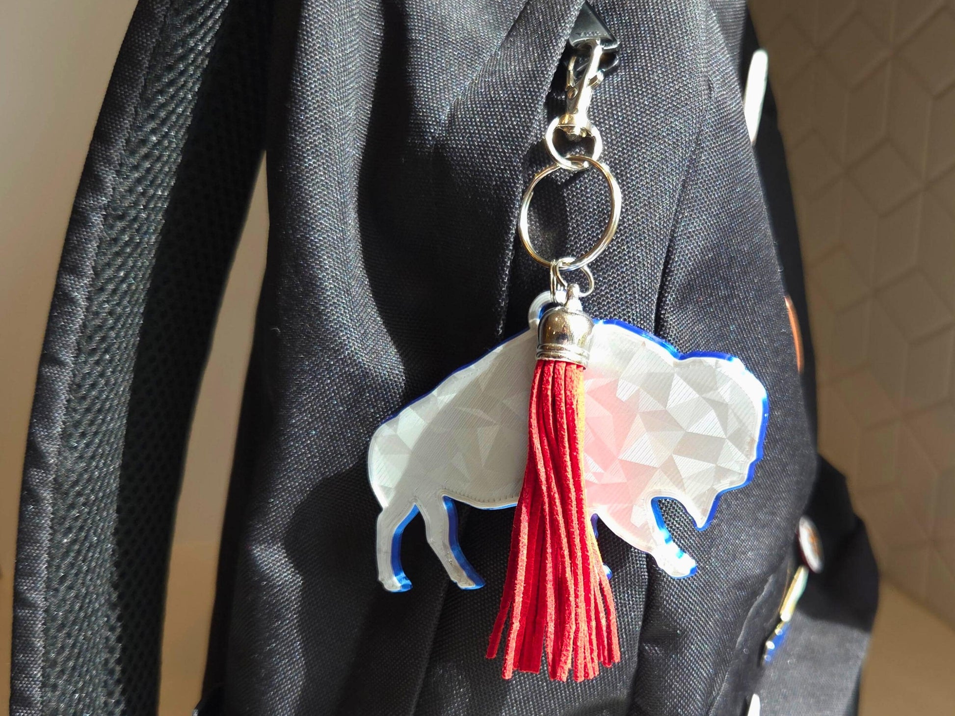 Buffalo football standing bison logo with geometric finish featured as a bakcpack charm in silver with a blue backing, and a red tassel for contrast.