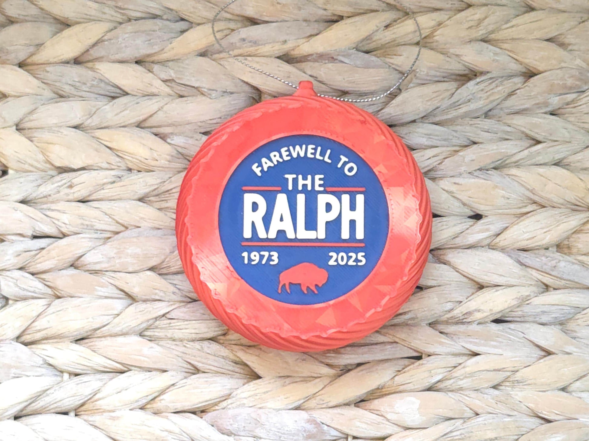 Close-up of a commemorative stadium ornament featuring raised white text and red accents, attached to a silver loop. The ornament is styled on a white dish with woven background texture.