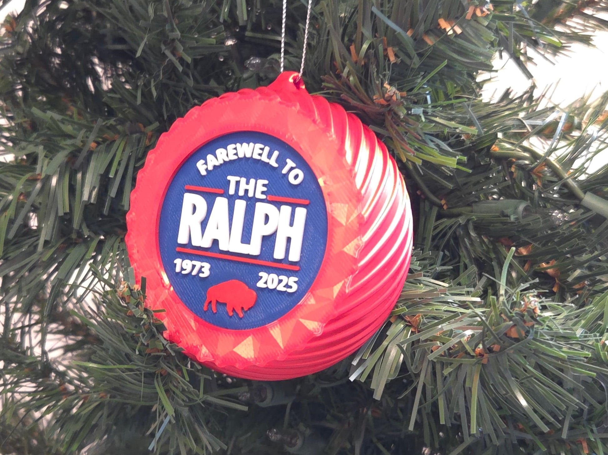 A 4" tall blue Christmas ornament with the words “Farewell to The Ralph 1973–2025” in bold white letters, a red buffalo graphic, and a silver hanging loop. Displayed hanging from a Christmas tree.