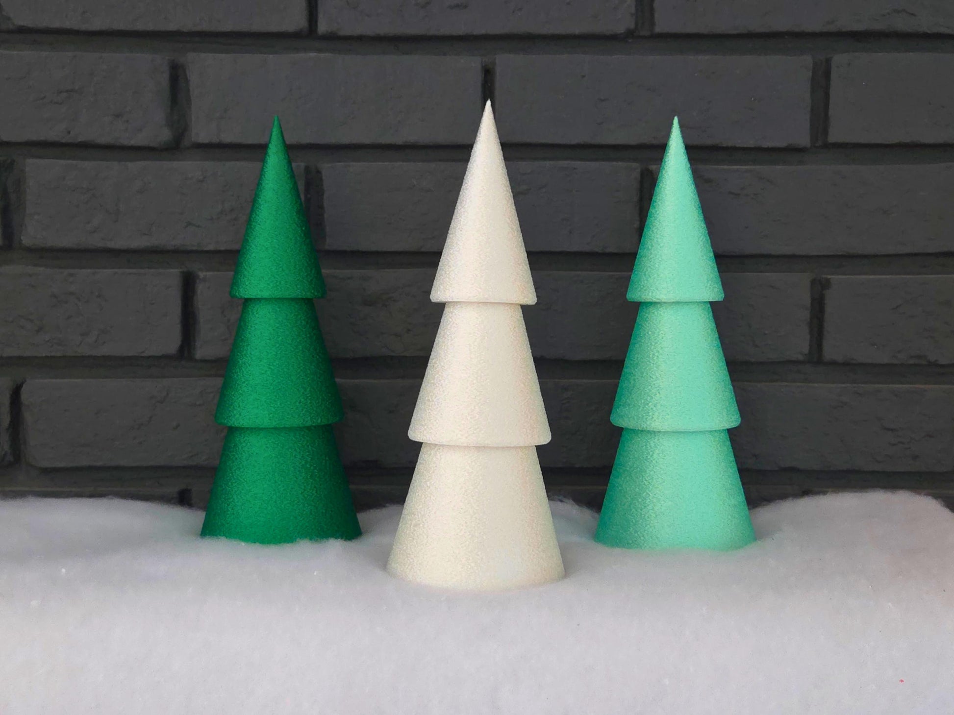 Set of three minimalist Christmas trees in dark green, light green and white pictured on a snowy backdrop with a black brick background.