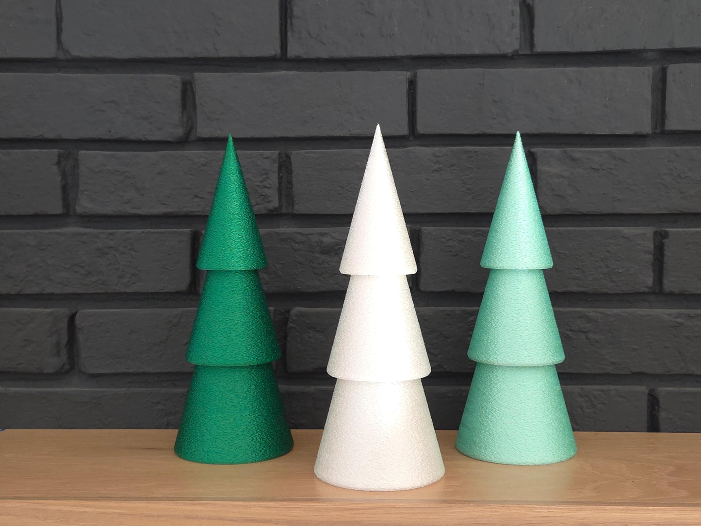 Set of three minimalist Christmas trees in dark green, light green and white pictured on a wooden shelf with a black brick background.