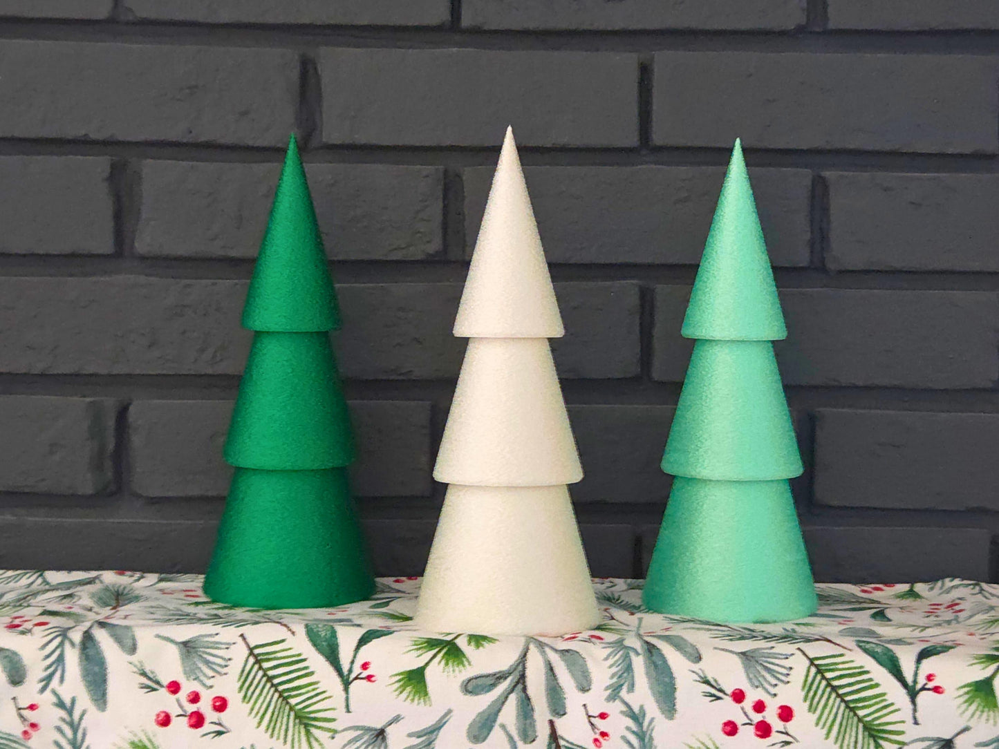 Set of three minimalist Christmas trees in dark green, light green and white pictured on holiday tablecloth with a black brick background.