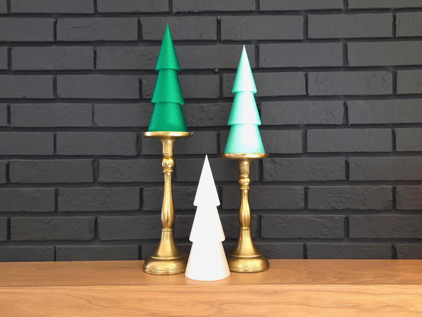 Set of three minimalist Christmas trees in dark green, light green and white pictured on a wooden shelf and with some candleabras with a black brick background.