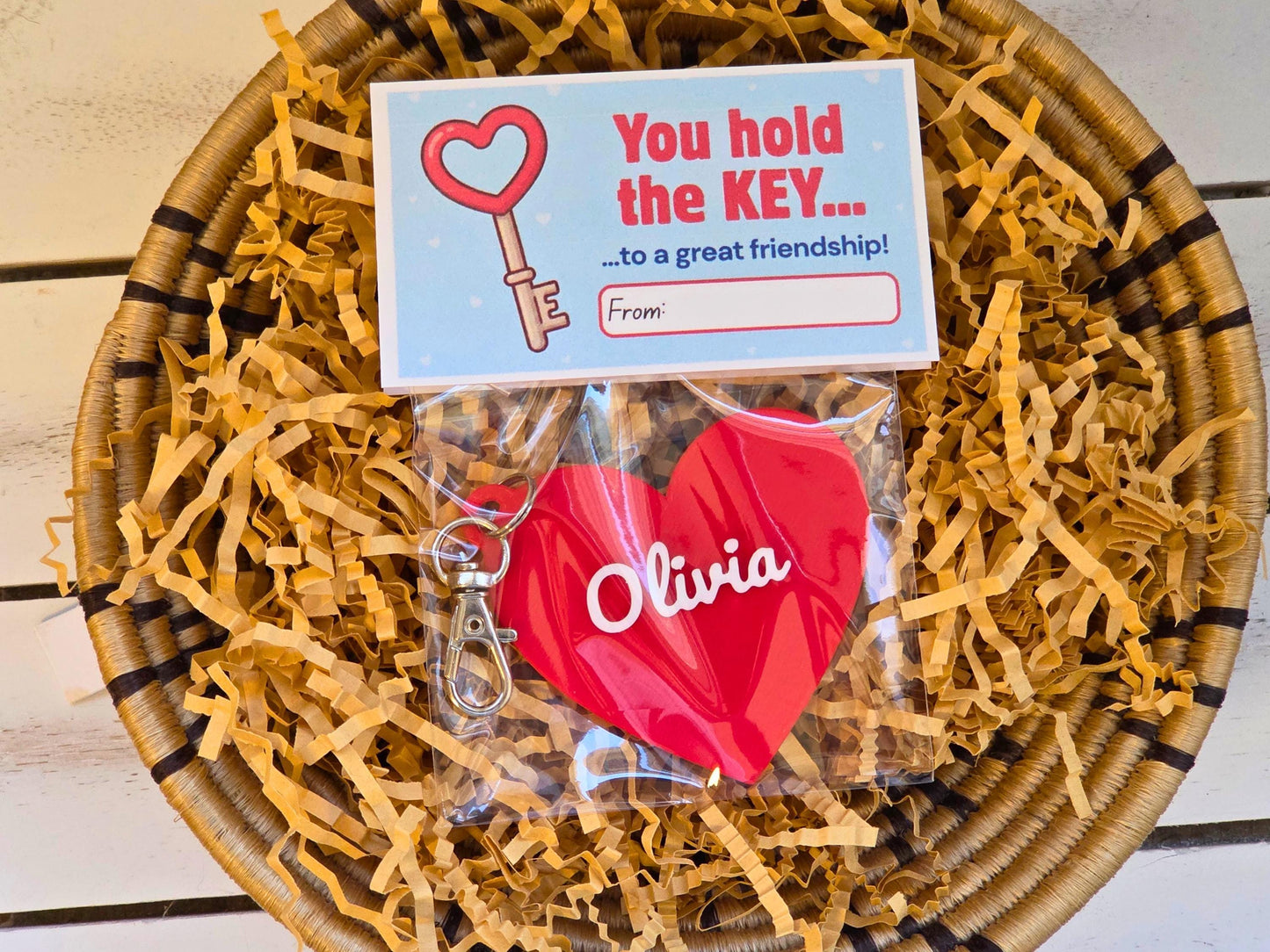 The front of the kid's Valentine gift is pictured in kraft paper on a wooden background. Each is a personalized heart keychain, which could also be used as a zipper pull or bag charm. It features the child's name, in this case the name Olivia. At the top of the bag, the bag topper serves as the Valentine with the message "You hold the key to a great friendship" with space for the child to write who the gift is from. On the back it says "Happy Valentine's Day"