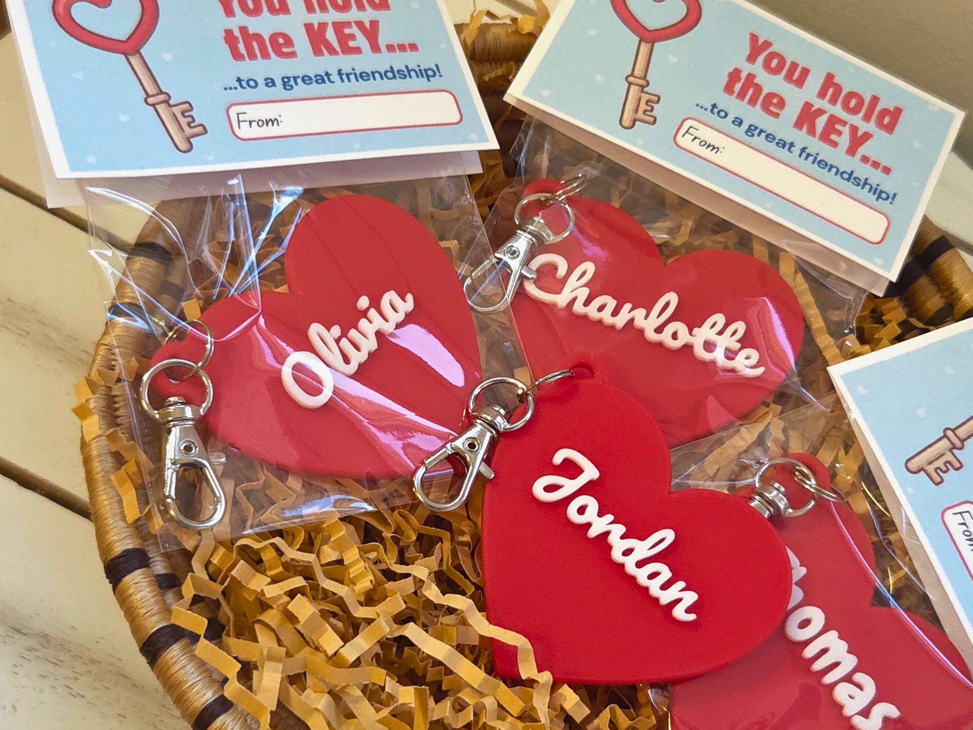 Classroom valentines are pictured in a dish of kraft paper on a wooden background. Each is a personalized heart keychain, which could also be used as a zipper pull or bag charm. It features the child's name, in this case the name Olivia, Charlotte, Jordan. At the top of the bag, the bag topper serves as the Valentine with the message "You hold the key to a great friendship" with space for the child to write who the gift is from. On the back it says "Happy Valentine's Day&q