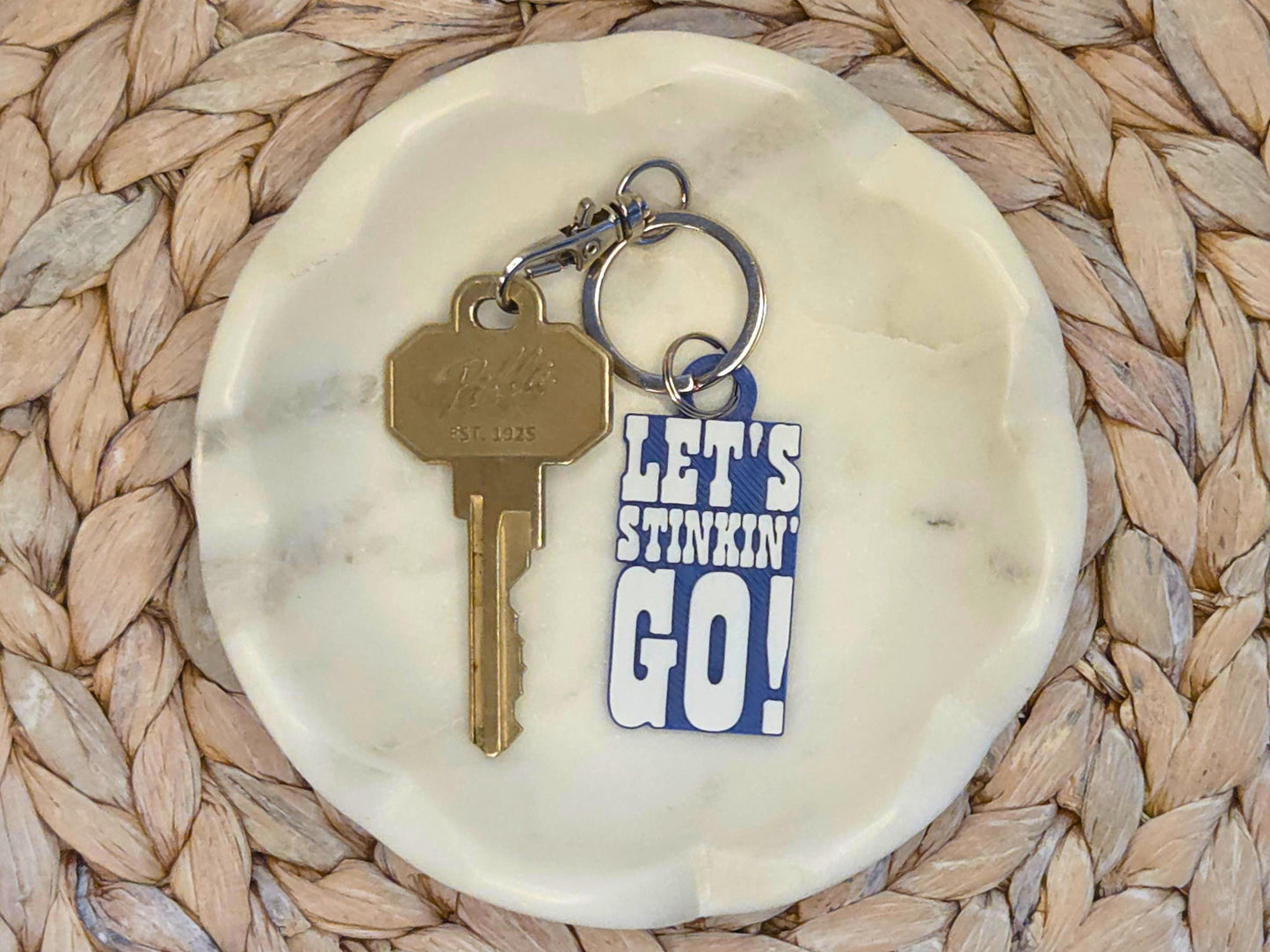Indianpolis Quarterback Keychain featuring mic'd up language Let's Stinkin' Go in white text on a blue background. Pictured in a white dish with a key for scale, on a woven background.