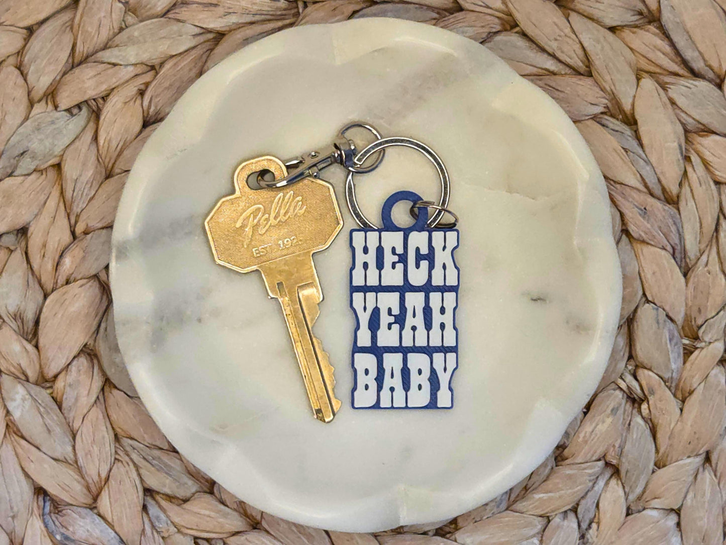 Indianpolis Quarterback Keychain featuring mic'd up language Heck Yeah Baby in white text on a blue background. Pictured in a white dish with a key for scale, on a woven background.