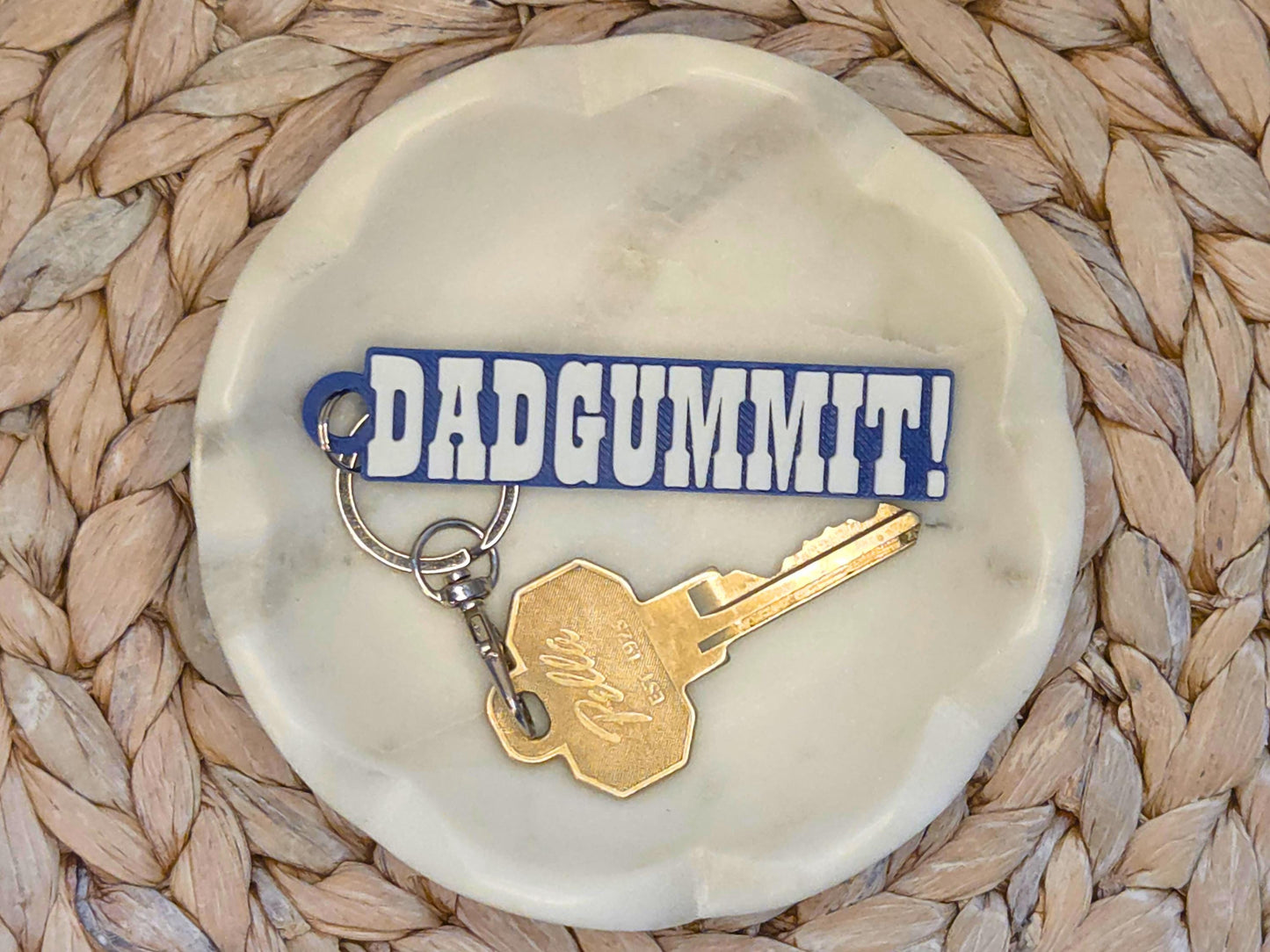 Indianpolis Quarterback Keychain featuring mic'd up language Dadgummit! in white text on a blue background. Pictured in a white dish with a key for scale, on a woven background.