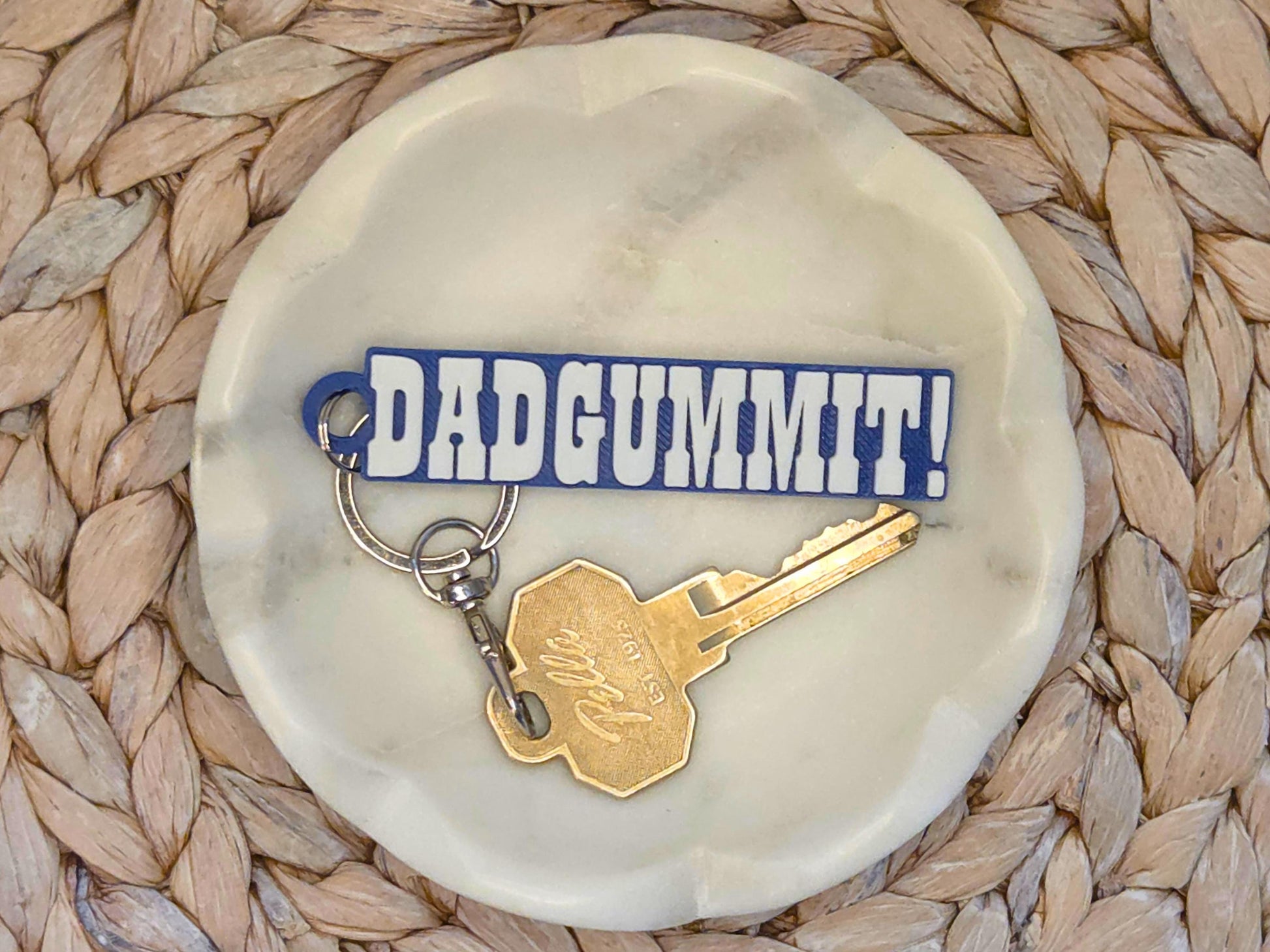 Indianpolis Quarterback Keychain featuring mic'd up language Dadgummit! in white text on a blue background. Pictured in a white dish with a key for scale, on a woven background.