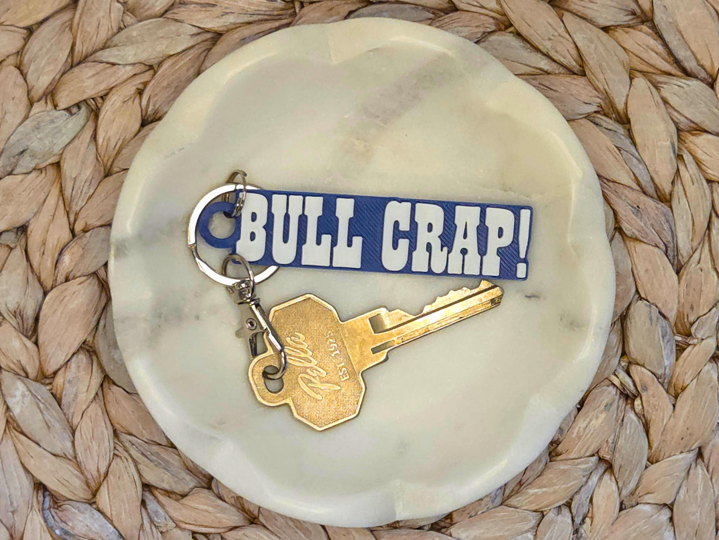 Indianpolis Quarterback Keychain featuring mic'd up language Bull Crap! in white text on a blue background. Pictured in a white dish with a key for scale, on a woven background.