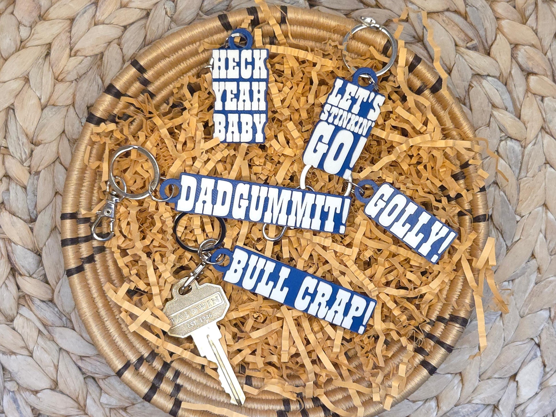 Set of five Indianpolis Quarterback Keychains featuring mic'd up language including Dadgummit, Let's Stinkin' Go, Golly!, Bull Crap!, Heck Yeah Baby in white text on a blue background. Pictured on kraft paper in a woven dish on a woven background.