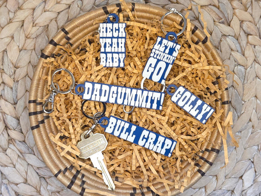 Set of five Indianpolis Quarterback Keychains featuring mic'd up language including Dadgummit, Let's Stinkin' Go, Golly!, Bull Crap!, Heck Yeah Baby in white text on a blue background. Pictured on kraft paper in a woven dish on a woven background.