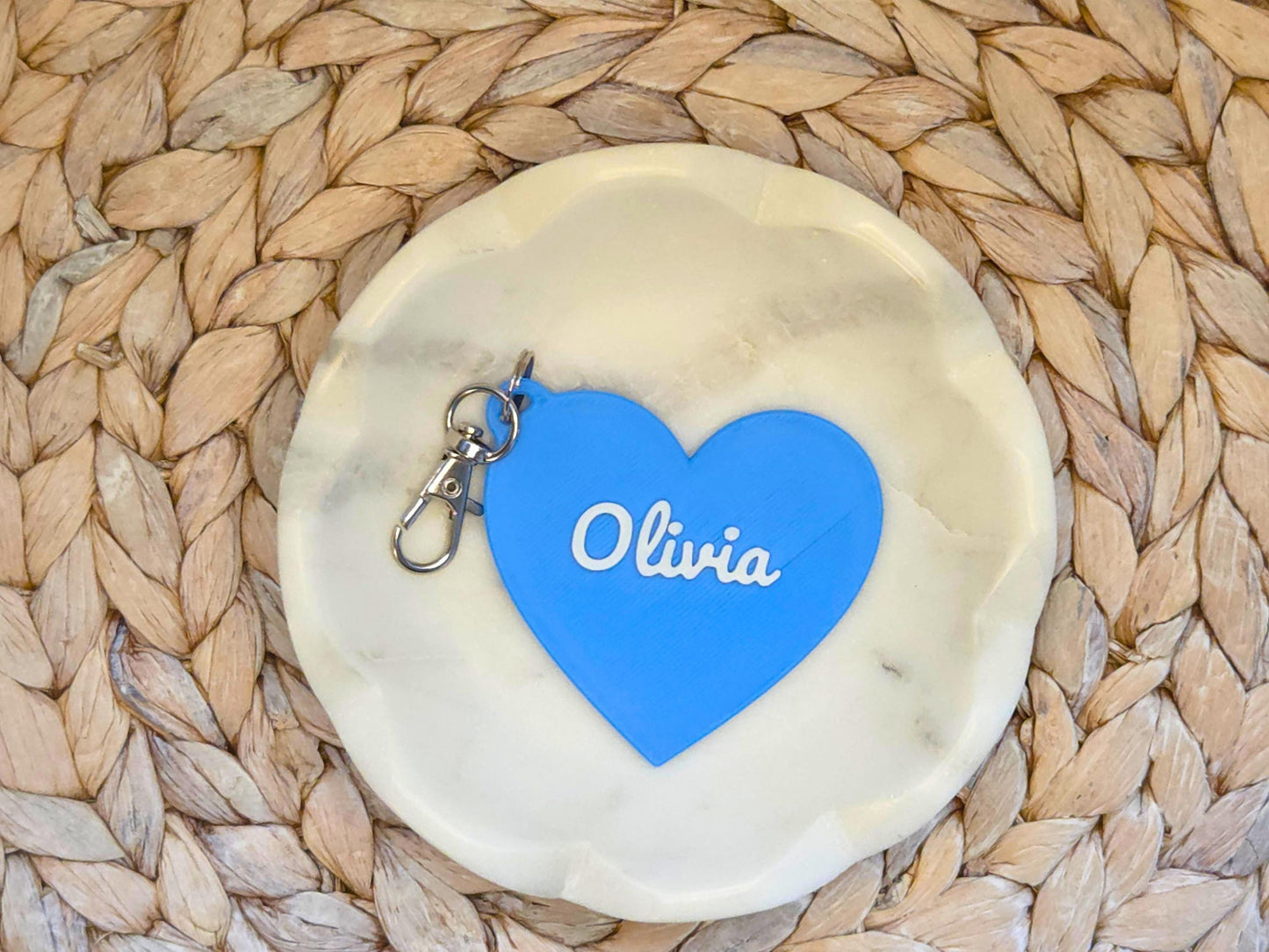 The front of the personalized heart keychain is pictured in blue in a white dish on a woven background. It can also be used as a zipper pull or bag charm. It features the child's name, in this case the name Olivia in script font.